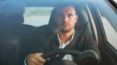 a car driver sits up and looks alert with wide eyes and an expression of concentration while he holds the wheel of a car.