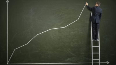 Man on a ladder drawing an increasing line on a chalk board symbolising a rising share price.