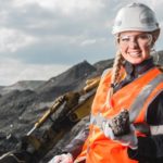 A female coal miner wearing a white hardhat and orange high-vis vest holds a lump of coal and smiles as the Whitehaven Coal share price rises today