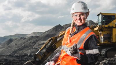 A female coal miner wearing a white hardhat and orange high-vis vest holds a lump of coal and smiles as the Whitehaven Coal share price rises today