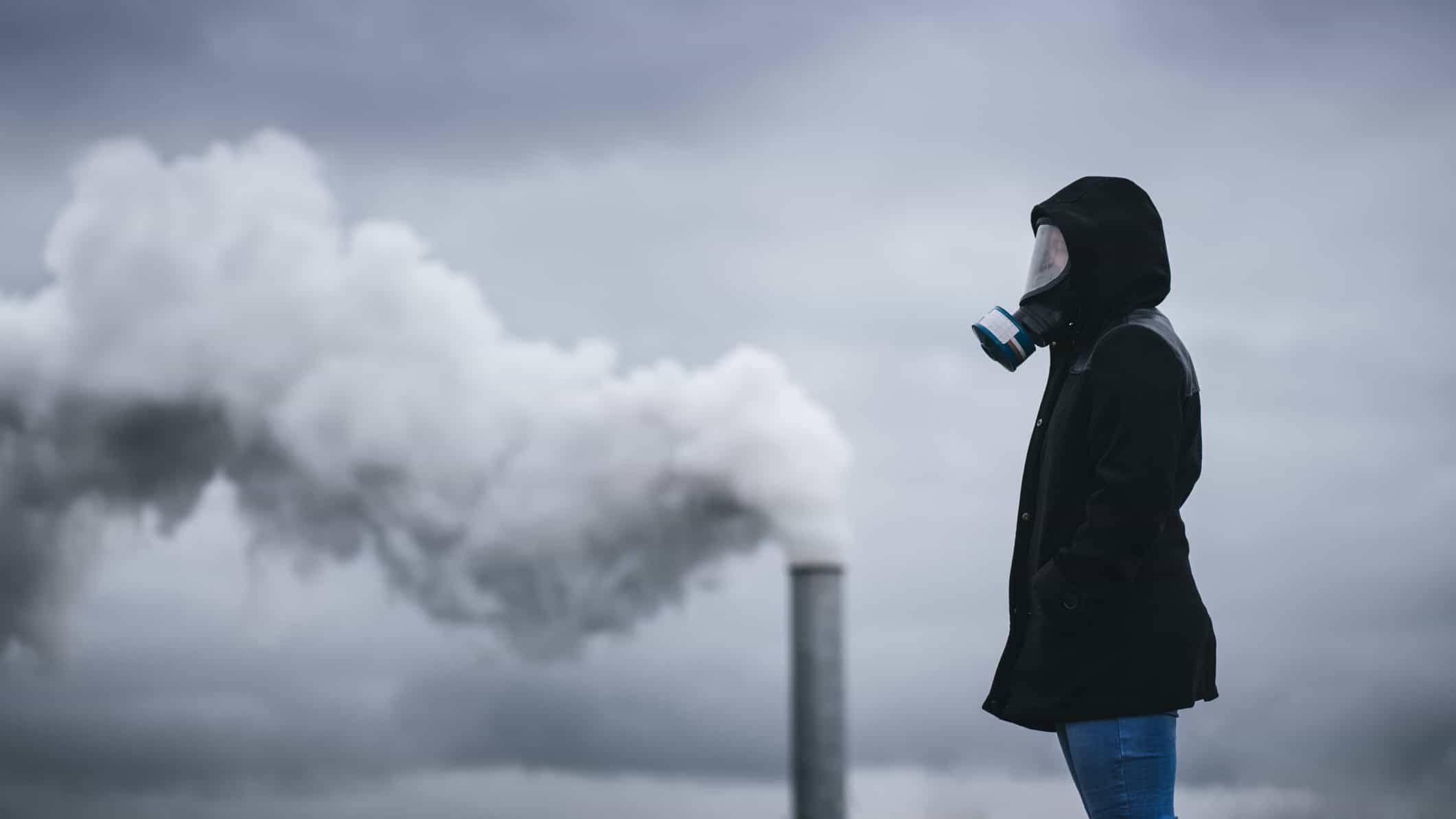 a person stands wearing a full old fashioned gas mask in the foreground of a coal fired smoke stack with smoke billowing into a grey sky and the person standing wearing a coat with hands in pockets.