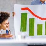 two people sitting at a desk look on in dismay as a colleague holds a chart with diminishing green bars topped with a jagged red line representing a stock market crash.
