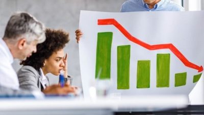 two people sitting at a desk look on in dismay as a colleague holds a chart with diminishing green bars topped with a jagged red line representing a stock market crash.