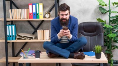 A young man in a blue suit sits on his desk cross-legged with his phone in his hand looking slightly crazed.