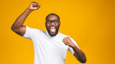 A man wearing glasses and a white t-shirt pumps his fists in the air looking excited and happy about the rising OBX share price