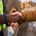 two hands shake in close up at the side of a mine. One party is wearing high visibility gear and there is earth and heavy moving equipment in the background.