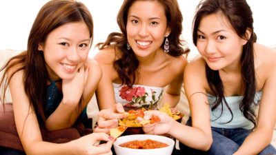 three young women smile as they hold up their loaded orn chips as they sit in front of a large bowl of dip.