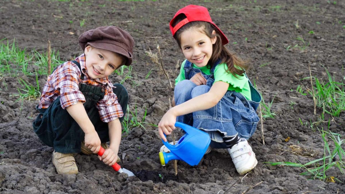 two children squat down in the dirt with gardening tools and a watering can wearing denim overalls and smiling very sweetly.