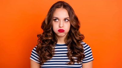 A young woman wearing a blue and white striped t-shirt blows air from her cheeks and looks up and to the side in a sign of disappointment after the ASX shares she owns went down today