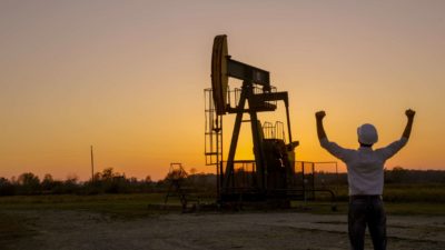 an oil worker holds his hands in the air in celebration in silhouette against a seitting sun with oil drilling equipment in the background.