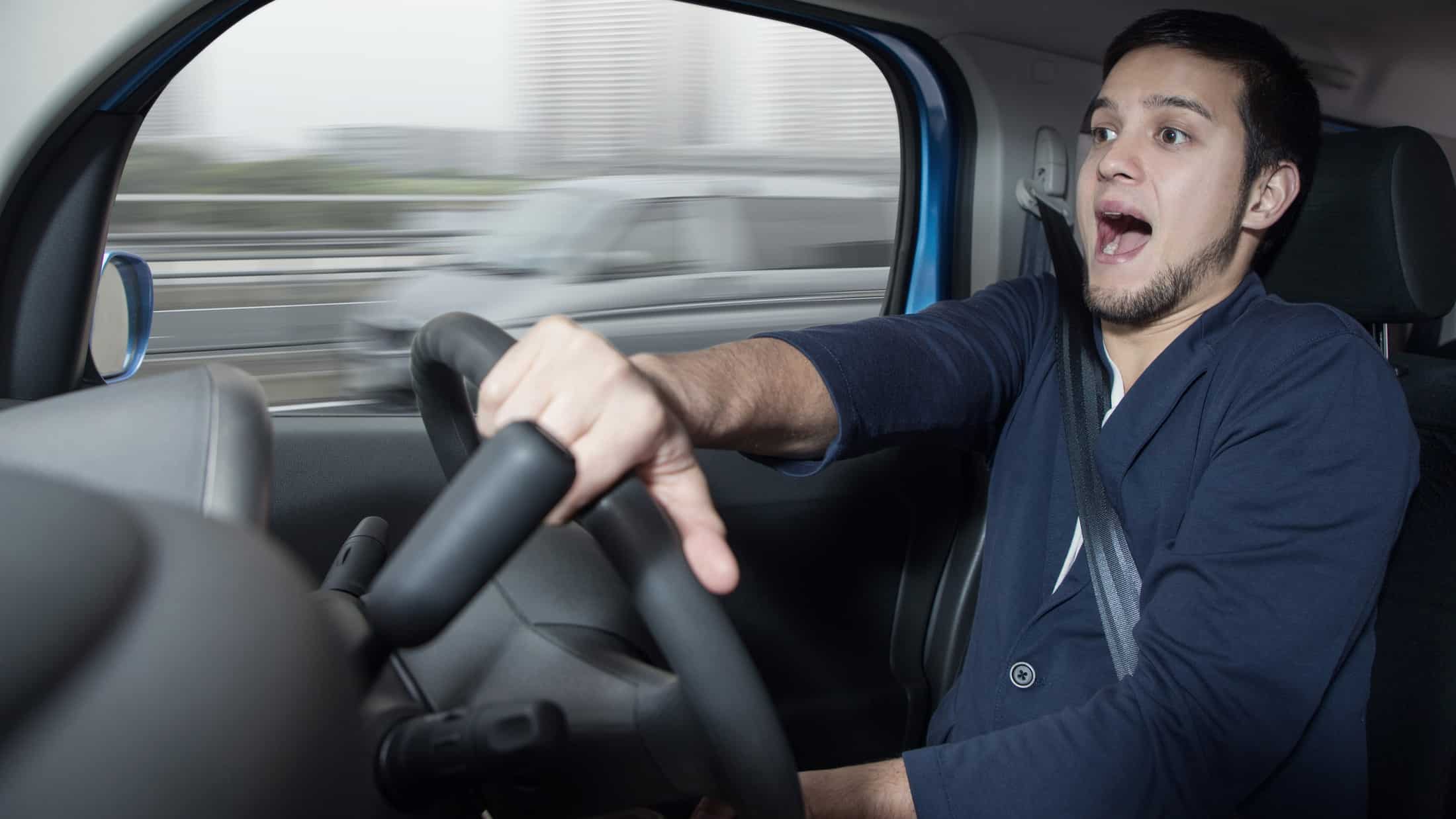 a man drives a car wearing a seat belt with a startled, almost frightened look on his face as he clutches the steering wheel as though he is having a wild ride in his car.