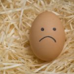 An egg with an unhappy face drawn on it lying on a bed of straw.