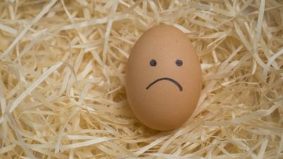 An egg with an unhappy face drawn on it lying on a bed of straw.