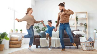 A husband and wife dance with their young daughter in their lounge room.
