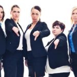 A group of five women in business attire stand side by side with unhappy looks on their faces and holding their thumbs down.