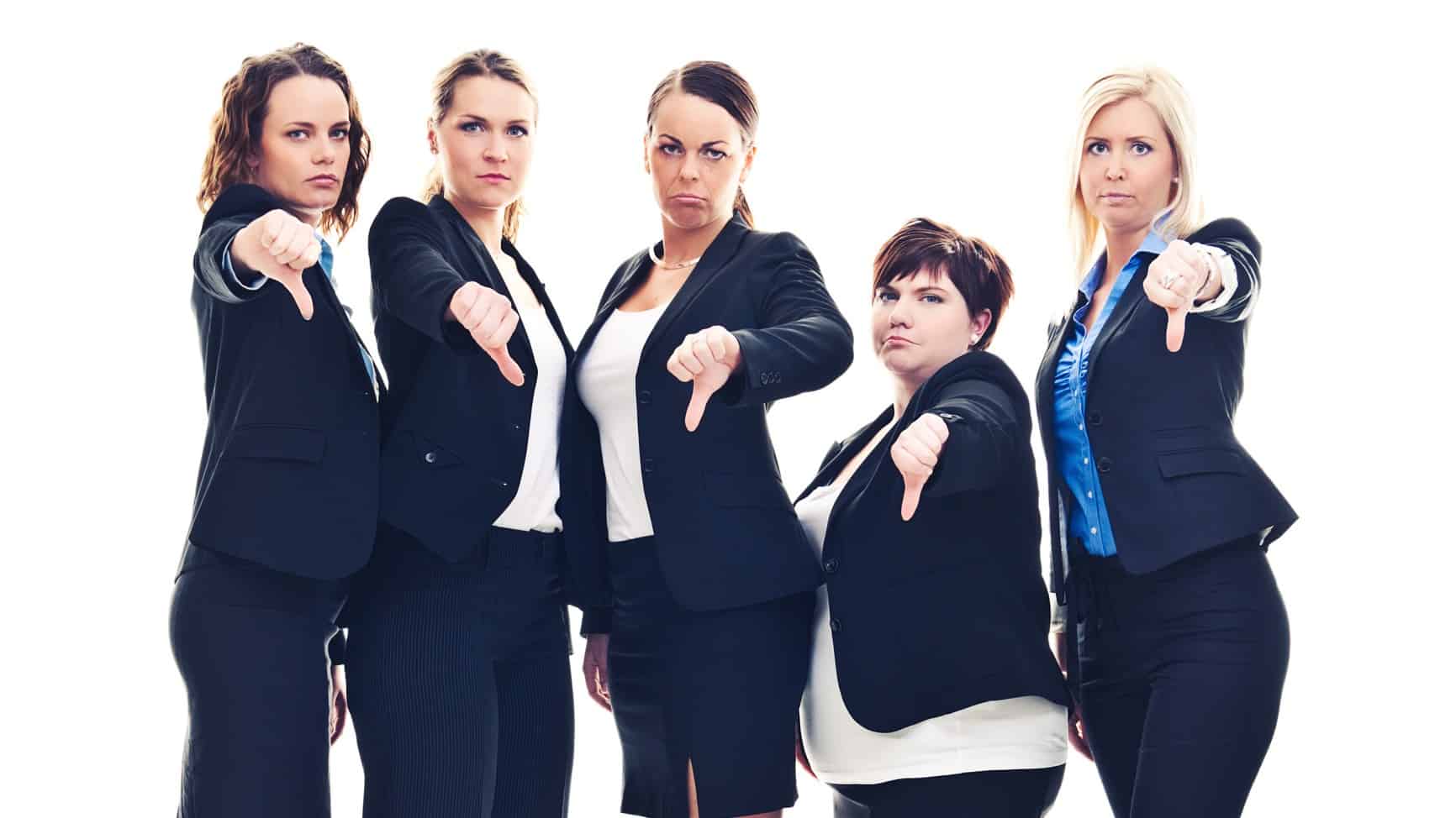 a group of five women in business attire stand side by side with unhappy looks on their faces and holding their thumbs down.