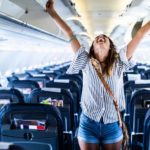 A woman on holiday stands with her arms outstretched joyously in an aeroplane cabin.
