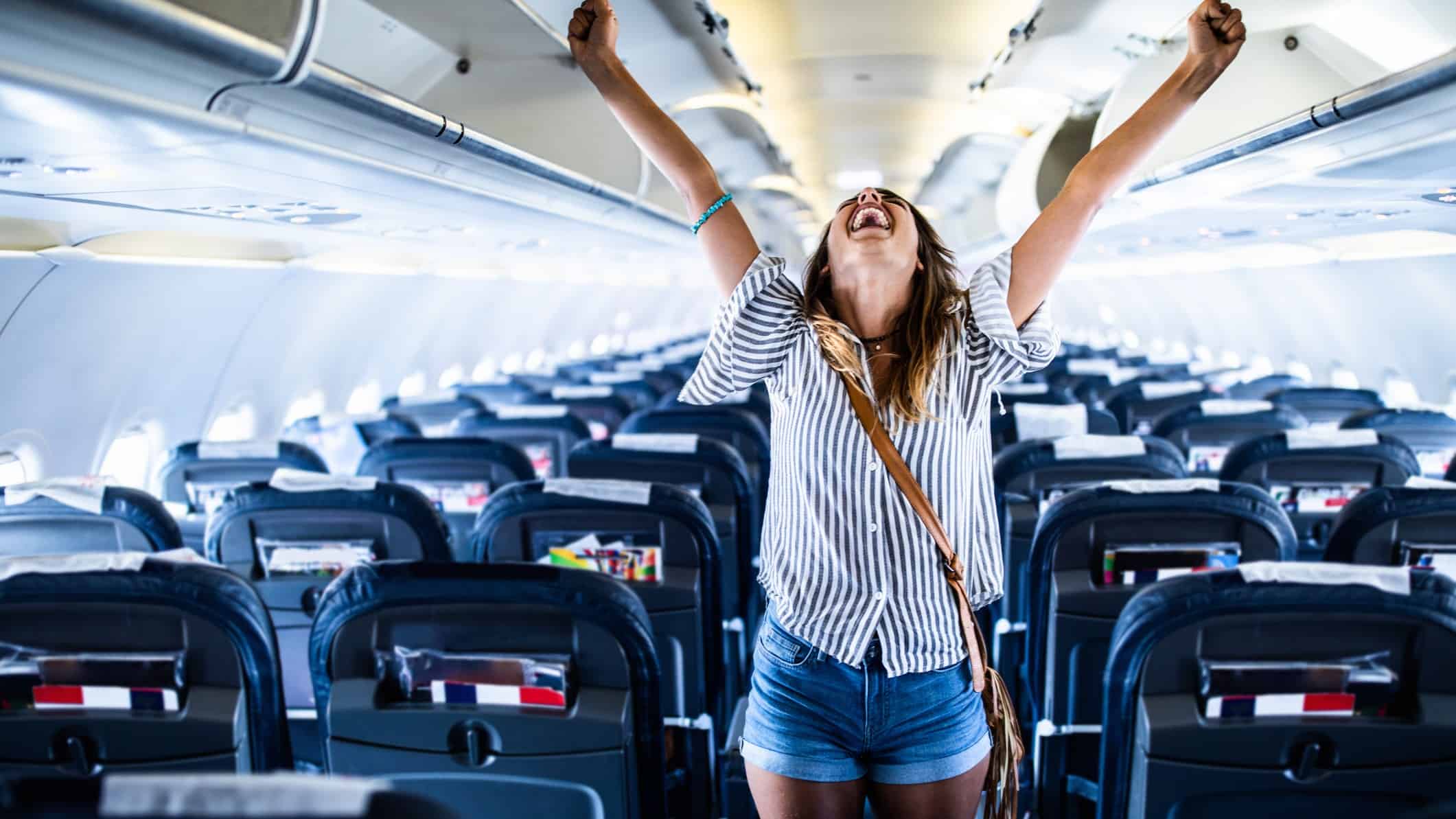 A woman wearing casual holiday attire stands with her head thrown back and her arms outstretched as if celebrating as she stands on board an empty plane with its rows of seats in the background.