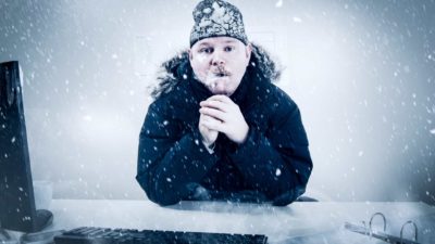 A businessperson sits at his desk in a cold office with snow and ice all around him and a frozen beard.