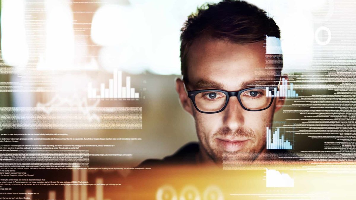 a man wearing spectacles has a satisfied look on his face as he appears within a graphic image of graphs, computer code and technology related symbols while he concentrates on a computer screen