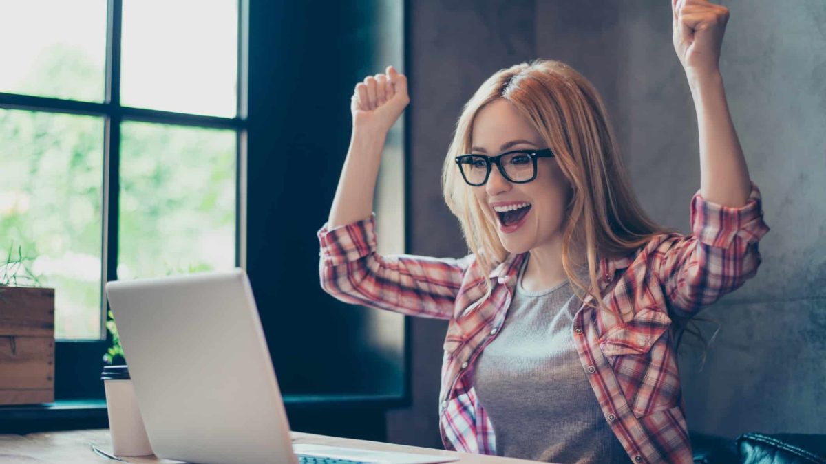 a young woman raises her hands in joyful celebration as she sits at her computer in a home environment.