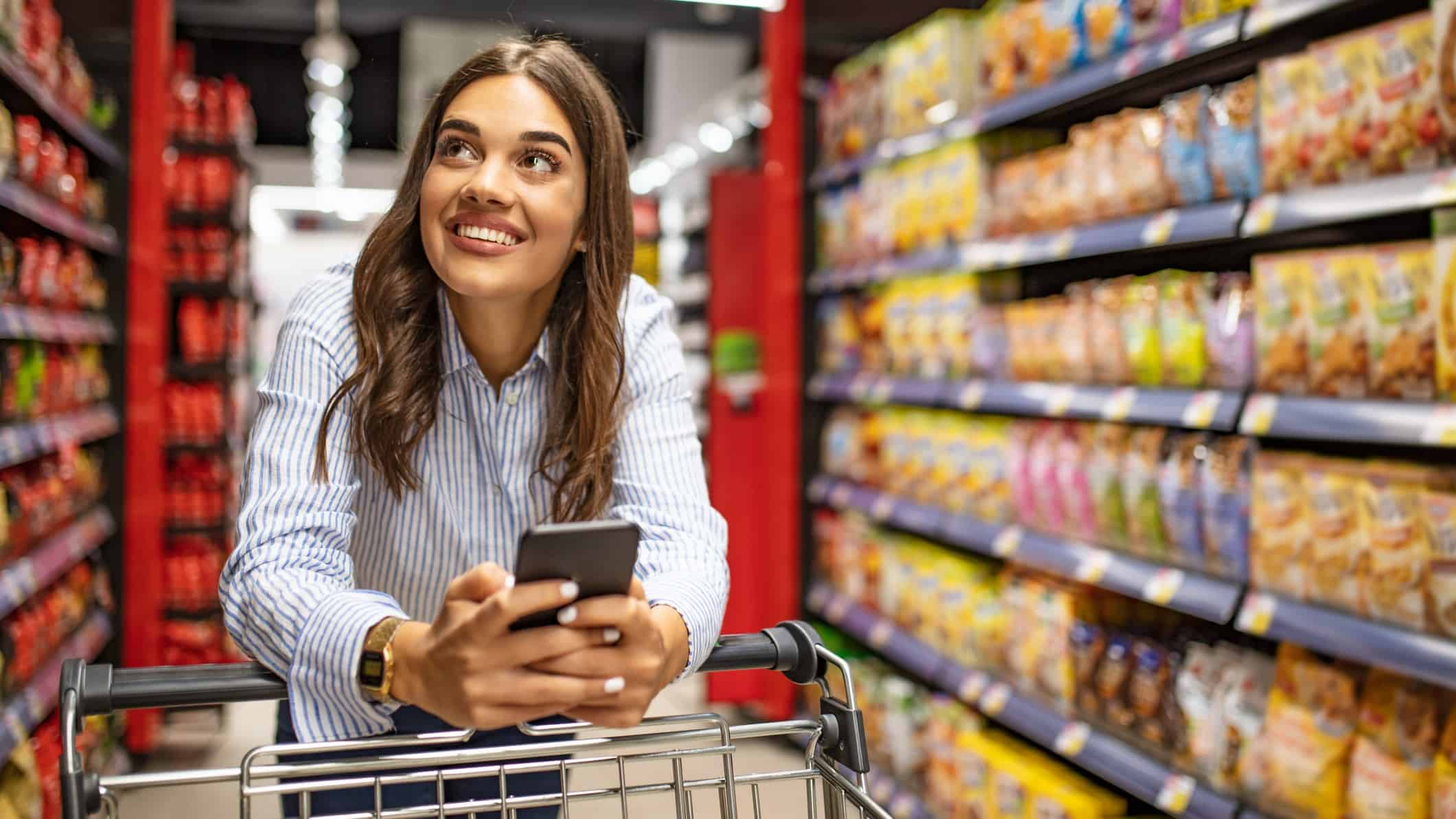a woman smiles widely as she leans on her trolley while making her way down a supermarket grocery aisle while holding her mobile telephone.