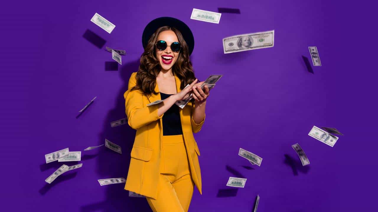 A laughing woman wearing a bright yellow suit, black glasses, and a black hat spins dollar bills out of her hands, reflecting dividend earnings.