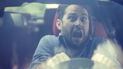 a wide-mouthed man looks scared as he grips the wheel of a car while driving in a murky environment.