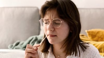 a woman winces as she inserts a home diagnostic test for COVID-19 up her nose with her sofa in the background.