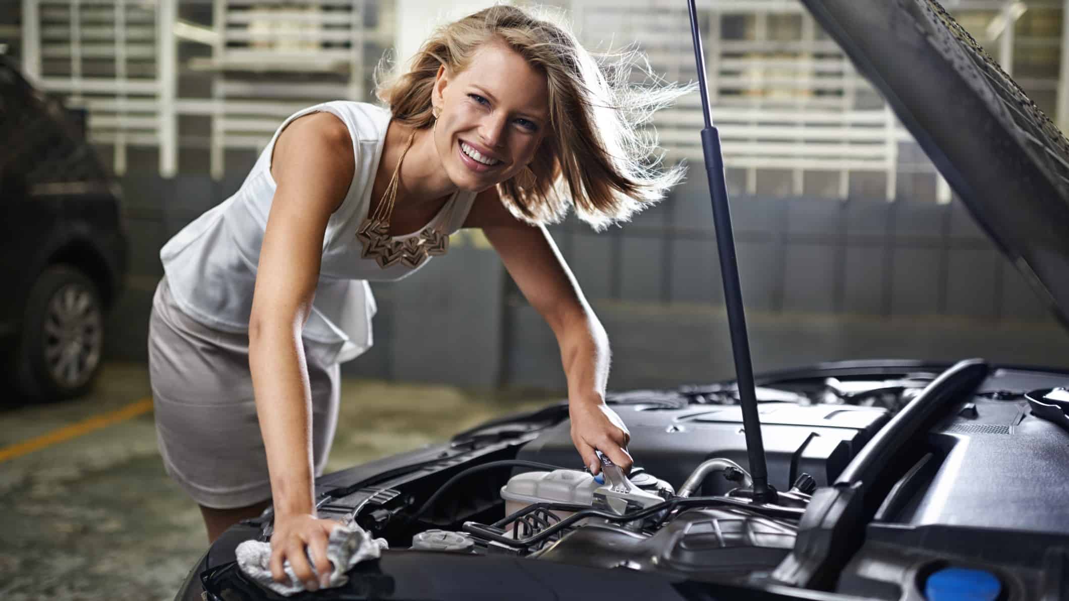 a smiling woman looks towards the camera as she tends to the engine under the lifted bonnet of her car.