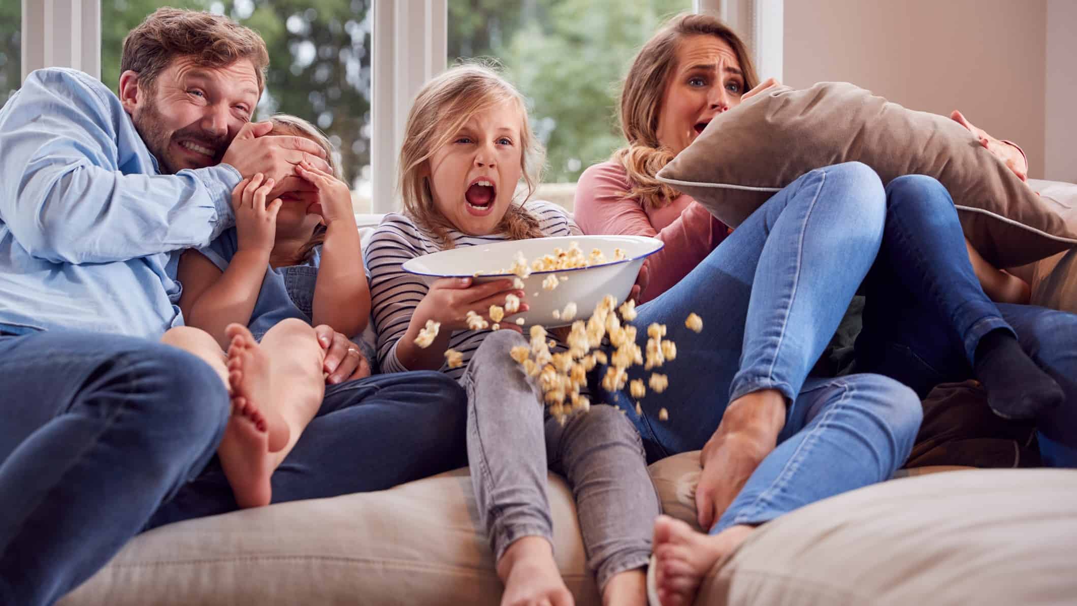 A family of three look scared as they watch a movie on the sofa with popcorn.