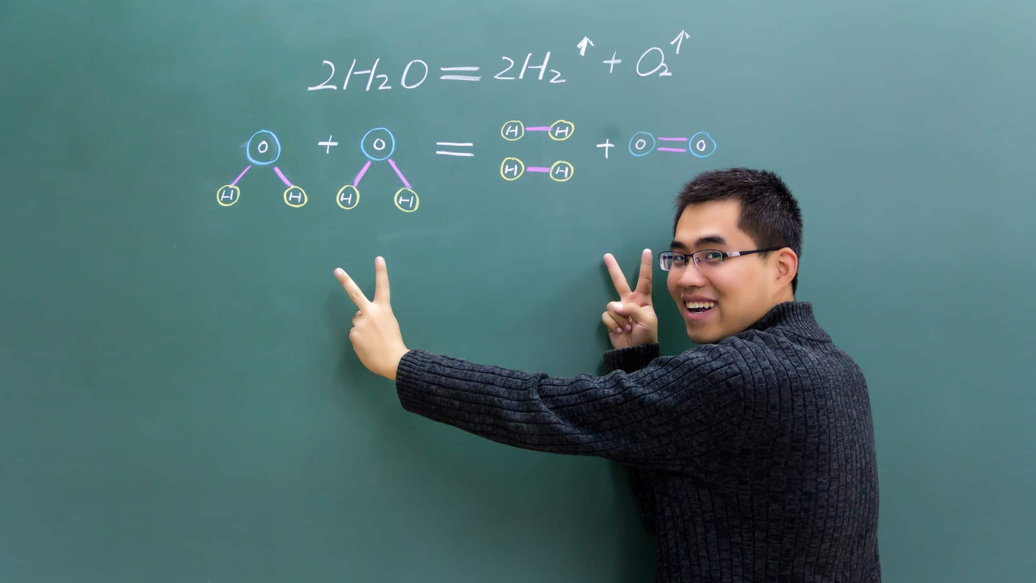 a man stands at a green blackboard where a scientific equation is written in chalk. He looks over his shoulder and holds two fingers of each hand in the air as he smiles, trying to illustrate the formation of hydrogen atoms.