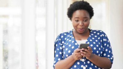 A young woman wearing a blue blouse with white polkadots holds her phone up with an intrigued and happy look on her face as she reads some news.