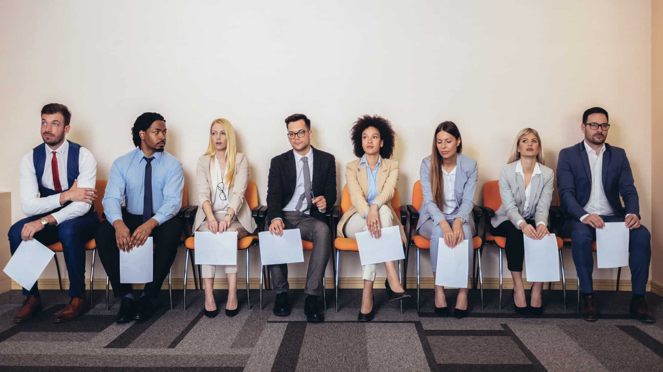 a line up of job interview candidates sit in chairs against a wall clutching CVs on paper in an office setting.