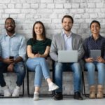 a line of job applicants sit on stools against a brick wall in an office environment, various holding laptops , devices and paper, as though waiting to be interviewed for a position.
