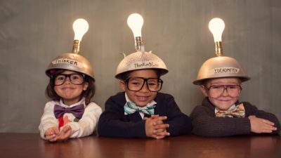 Three adorable children sit side by side at a table wearing upturned colanders on their heads fixed with shining light bulbs as they smile at the camera.