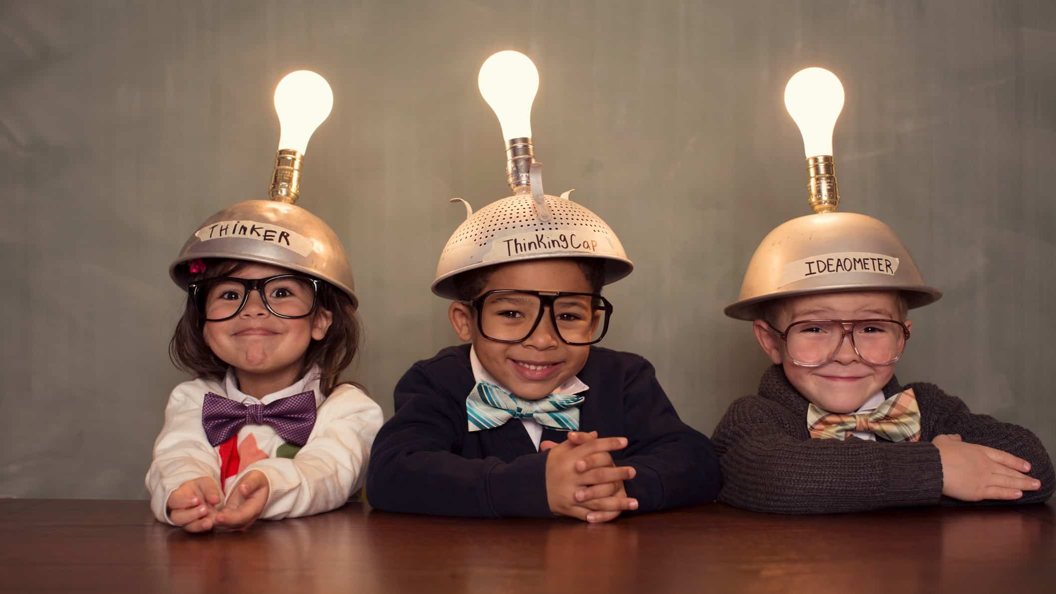 three adorable children sit side by side at a table wearing upturned colanders on their heads fixed with shining light bulbs as they smile cutely at the camera.