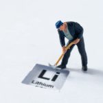 a miniature moulded model of a man bent over with a pick working stands behind a sign that has lithium's scientific abbreviation 'Li' with the word lithium underneath it against a sparse bland background.