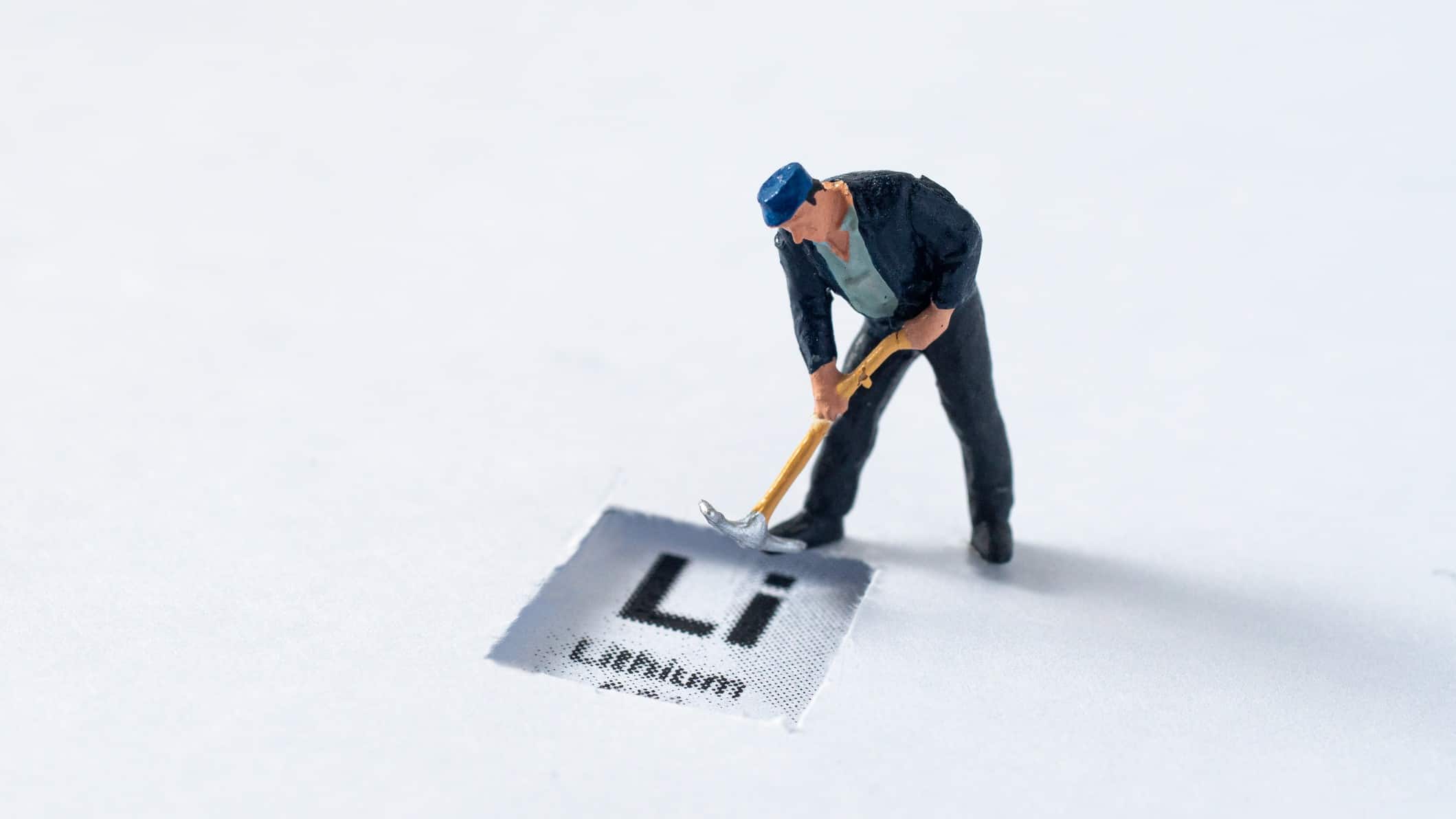a miniature moulded model of a man bent over with a pick working stands behind a sign that has lithium's scientific abbreviation 'Li' with the word lithium underneath it against a sparse bland background.