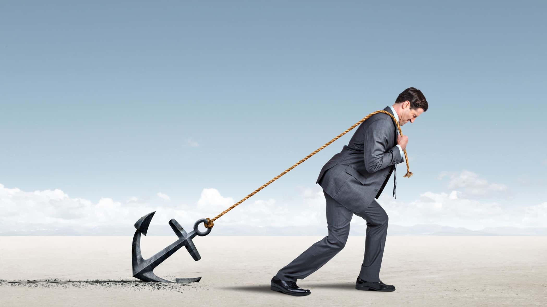 a businessman in a suit tries to forge ahead but is carrying a rope attached to a large anchor that is stuck in the ground against a background of muted sky and barren earth.