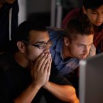 A group of people gather around a computer screen in rapt attention, one man holds his hands to cover his mouth as if in nervous anticipation of what news may come.