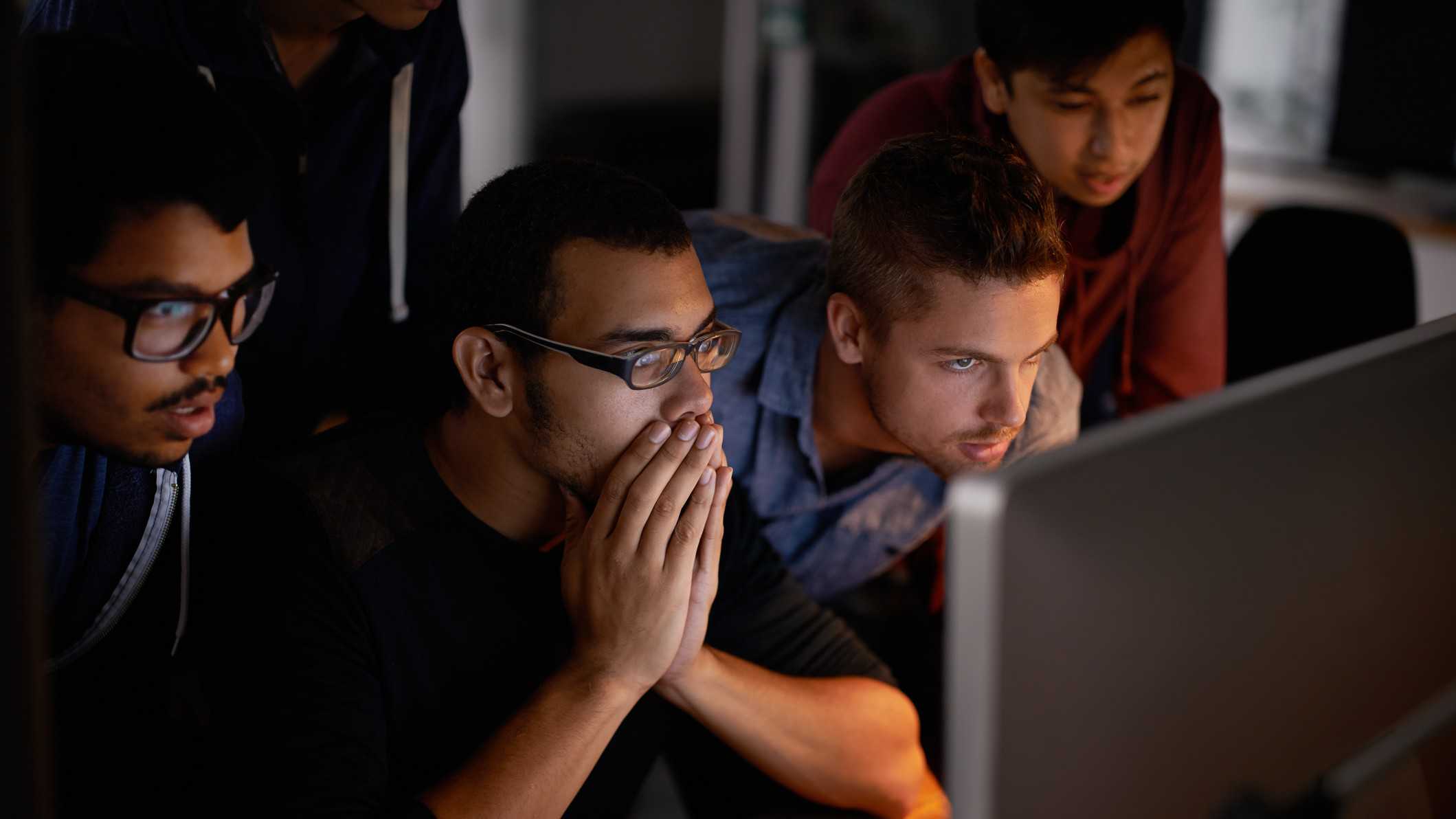 A group of people gather around a computer screen in rapt attention, one man holds his hands to cover his mouth as if in nervous anticipation of what news may come.