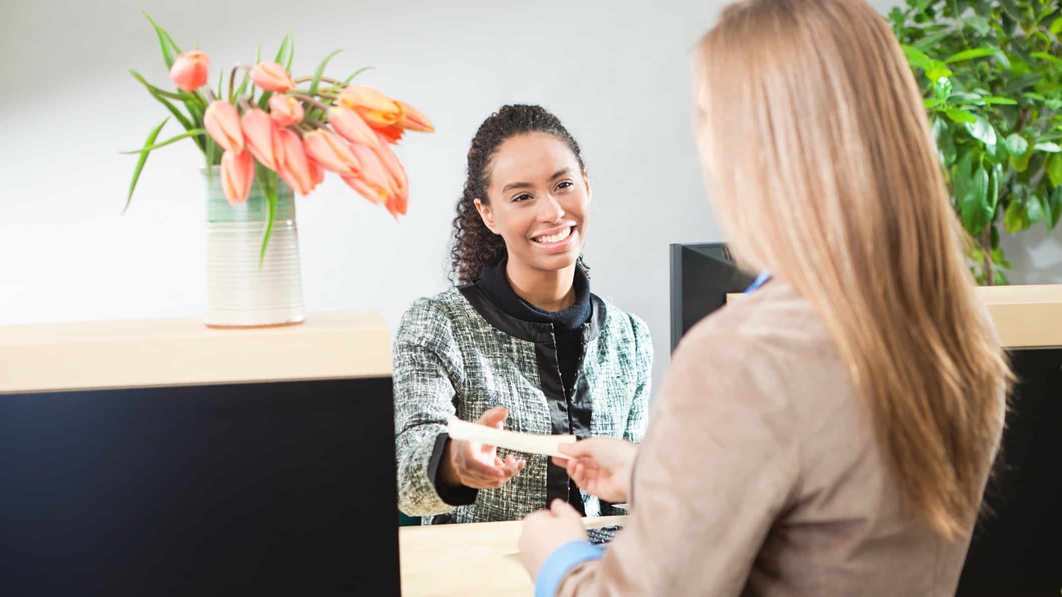 a female bank teller smiles warmly as she hands over a piece of paper to a female customer while a large vase of tulips rests on the bank counter.