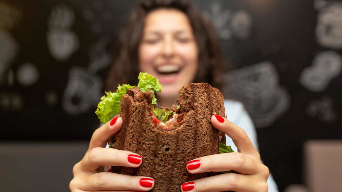 a wide-smiling woman holds her sandwich with a bite out of it towards the camera.
