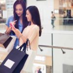 Two women are glamourously dressed in a shopping mall carrying designer shopping bags and looking excitedly at something on a mobile phone.