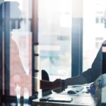 Two businessmen shake hands behind a window.