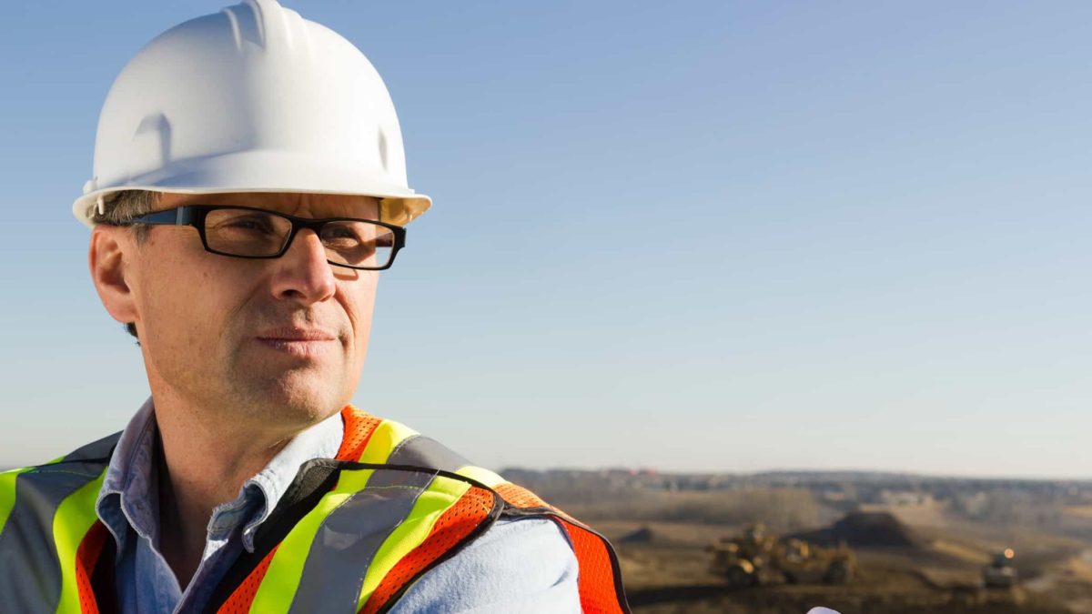 A man wearing a hard hat and high visibility vest looks out over a vast plain where heavy mining equipment can be seen in the background.