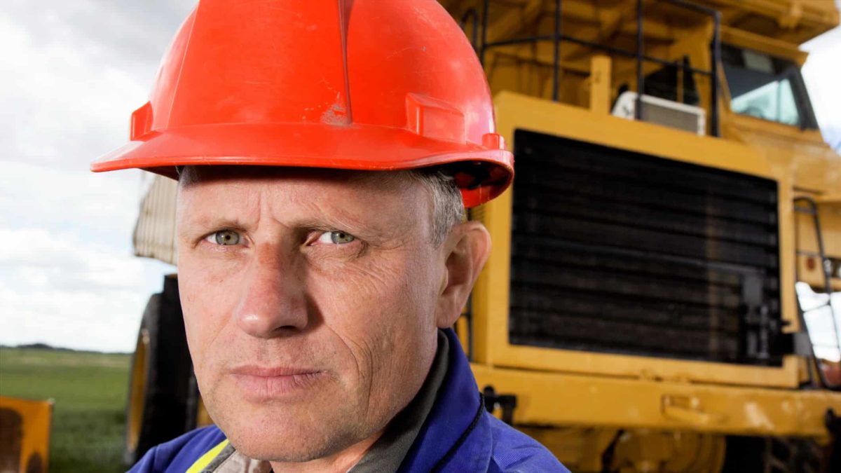 A man wearing a hard hat stands in front of heavy mining machinery with a serious look on his face.