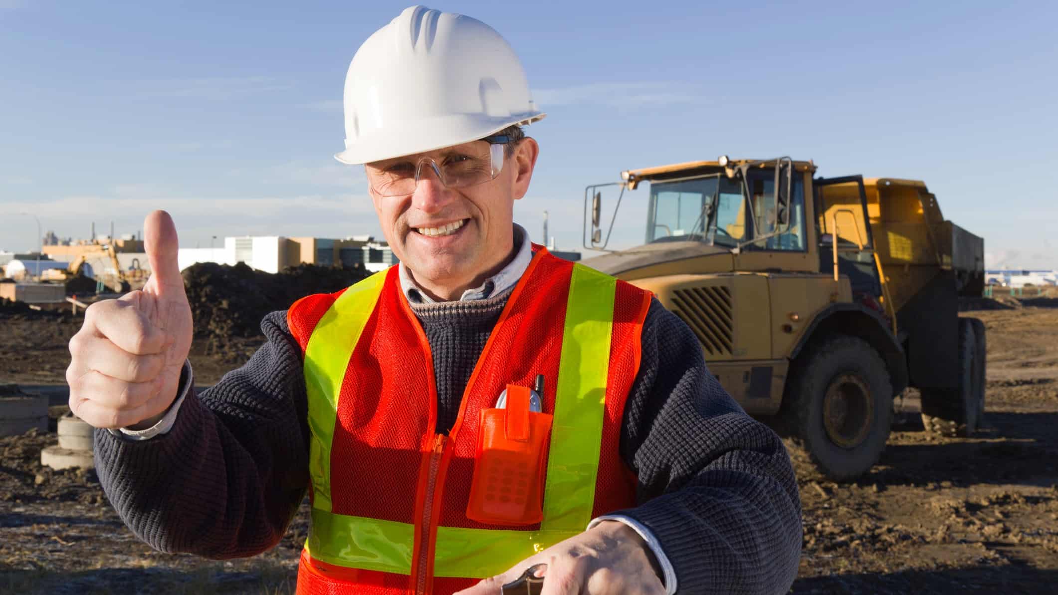 a man in a high visibility vest and hard hat holds a thumbs up at a mine site with heavy equipment in the background.
