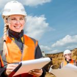 A female miner wearing a high vis vest and hard hard smiles and holds a clipboard while inspecting a mine site with a colleague.